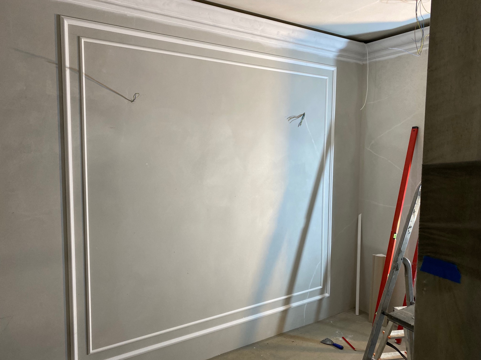 Ściana w trakcie wykańczania z zamontowanymi sztukateriami w kształcie prostokątnych ram, widoczne wystające kable elektryczne i drabina malarska.