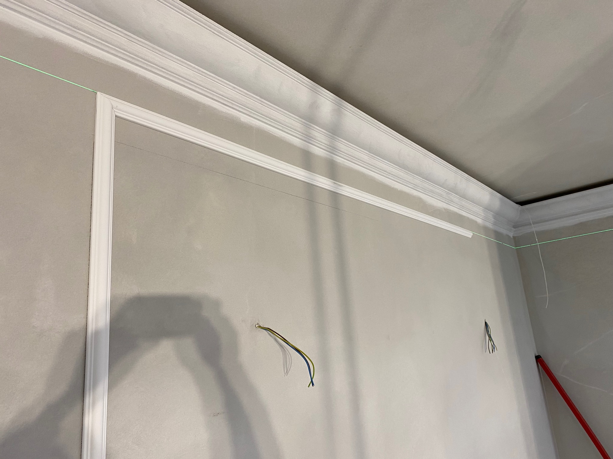 Ściana w trakcie wykańczania z dekoracyjnymi listwami przysufitowymi i ściennymi, widoczne przewody elektryczne wystające ze ściany oraz linia lasera wyznaczająca poziom.