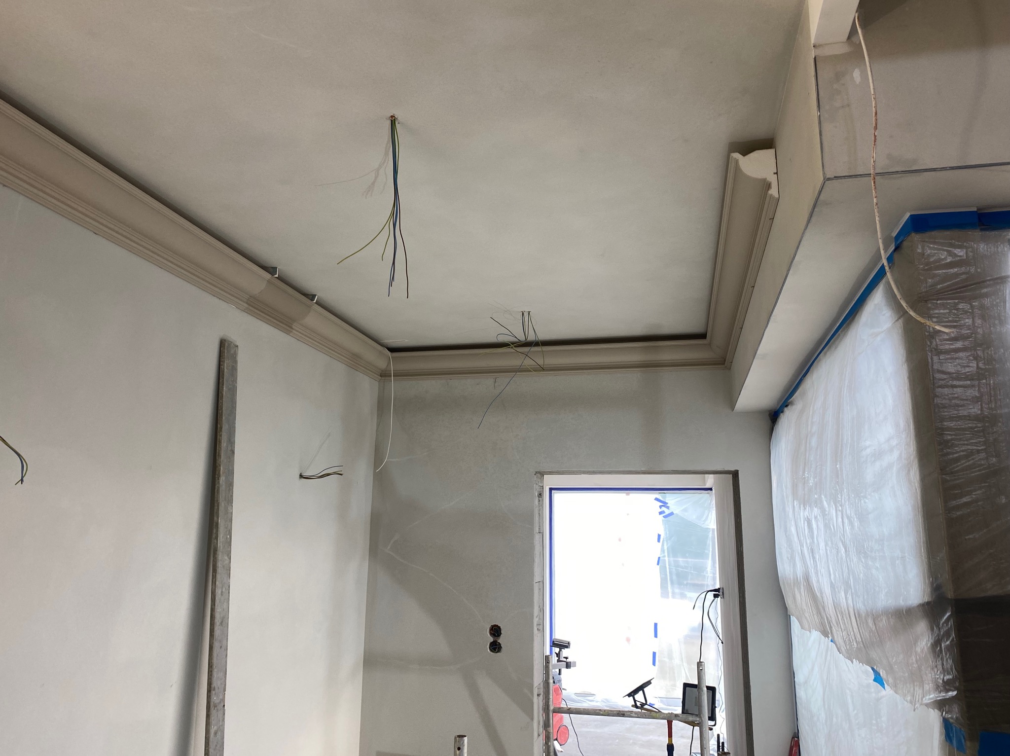 Surowe wnętrze w trakcie wykańczania: ściany i sufit po gładziowaniu, z widocznymi przewodami elektrycznymi, listwami dekoracyjnymi pod sufitem oraz zabezpieczonym folią oknem.