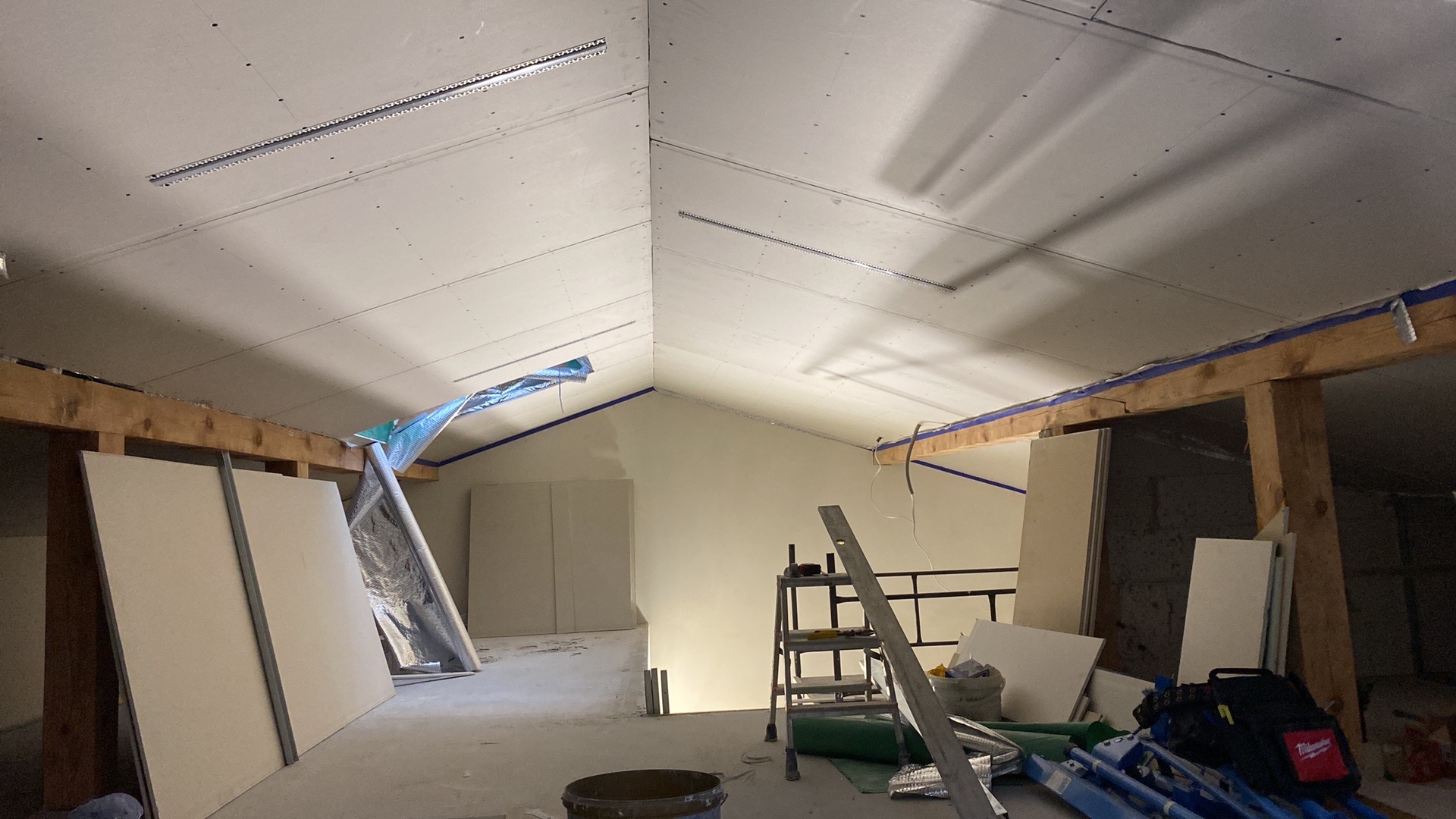 Poddasze w trakcie wykańczania: widoczne płyty kartonowo-gipsowe na suficie, drewniane belki stropowe zabezpieczone taśmą malarską, drabina, wiadro i narzędzia budowlane w nieładzie na podłodze.