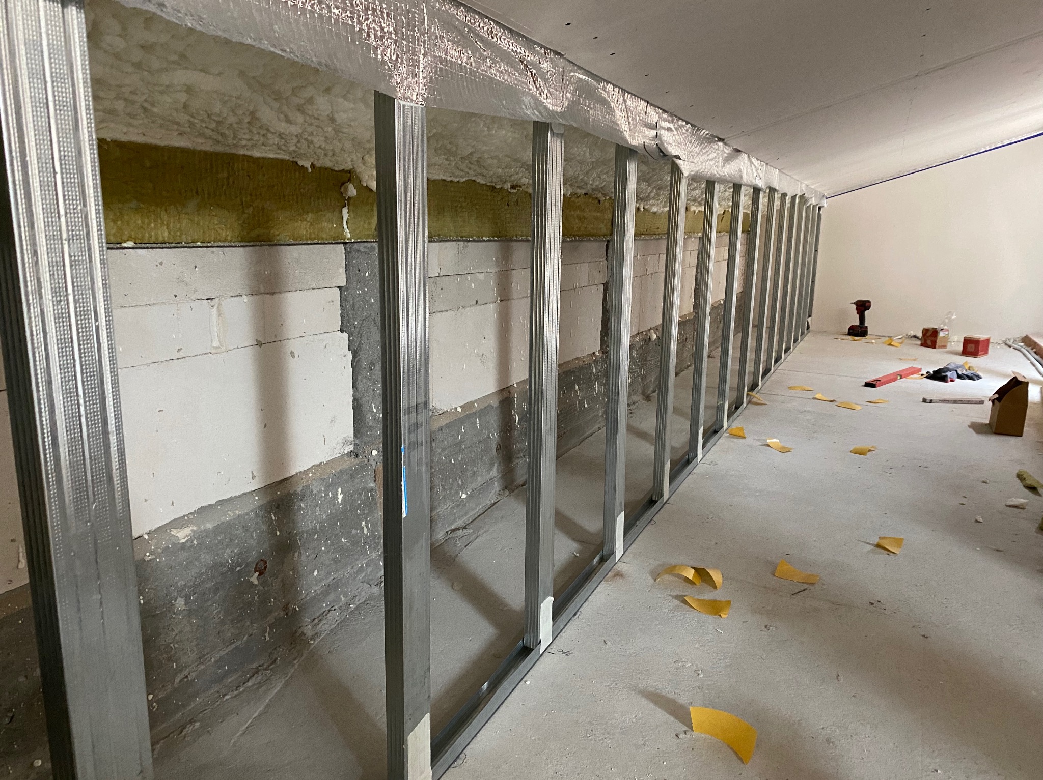 Montaż ściany działowej z płyt karton-gips: widoczna konstrukcja metalowa, ocieplenie wełną mineralną i pianką, częściowo wymurowana z bloczków betonowych, narzędzia i resztki taśmy na podłodze.