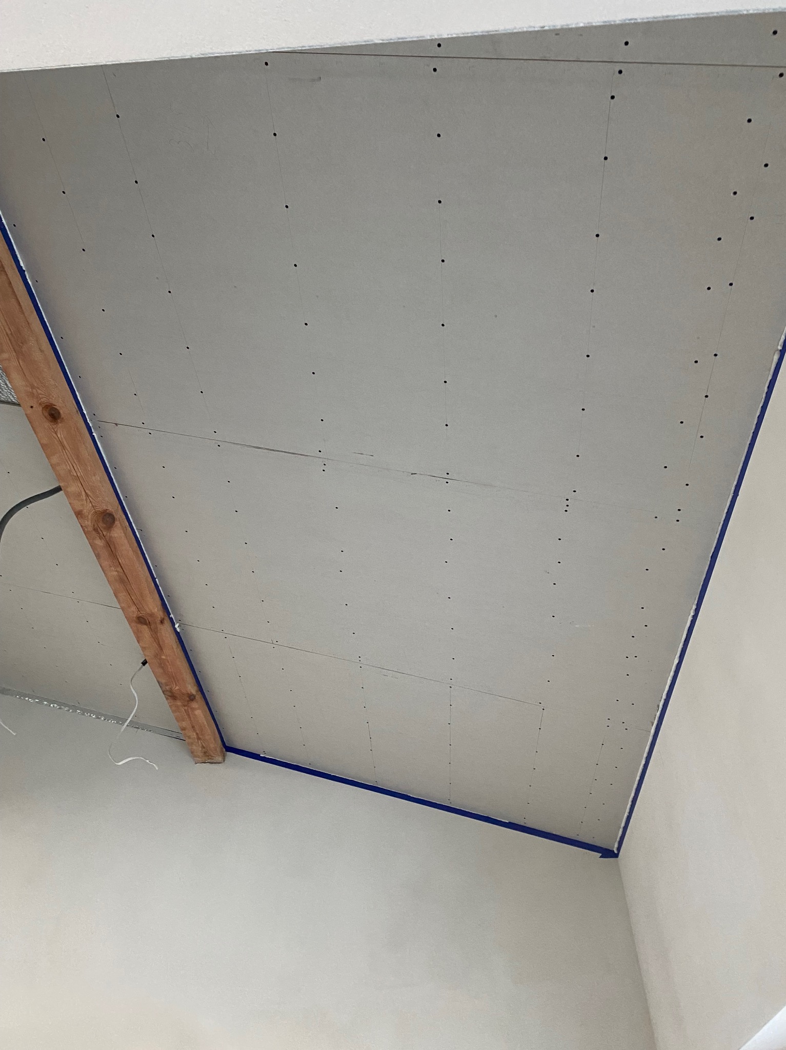 Montaż płyty gipsowo-kartonowej na suficie, widoczne wkręty w regularnych odstępach, drewniana belka konstrukcyjna z niebieską taśmą malarską na krawędziach.