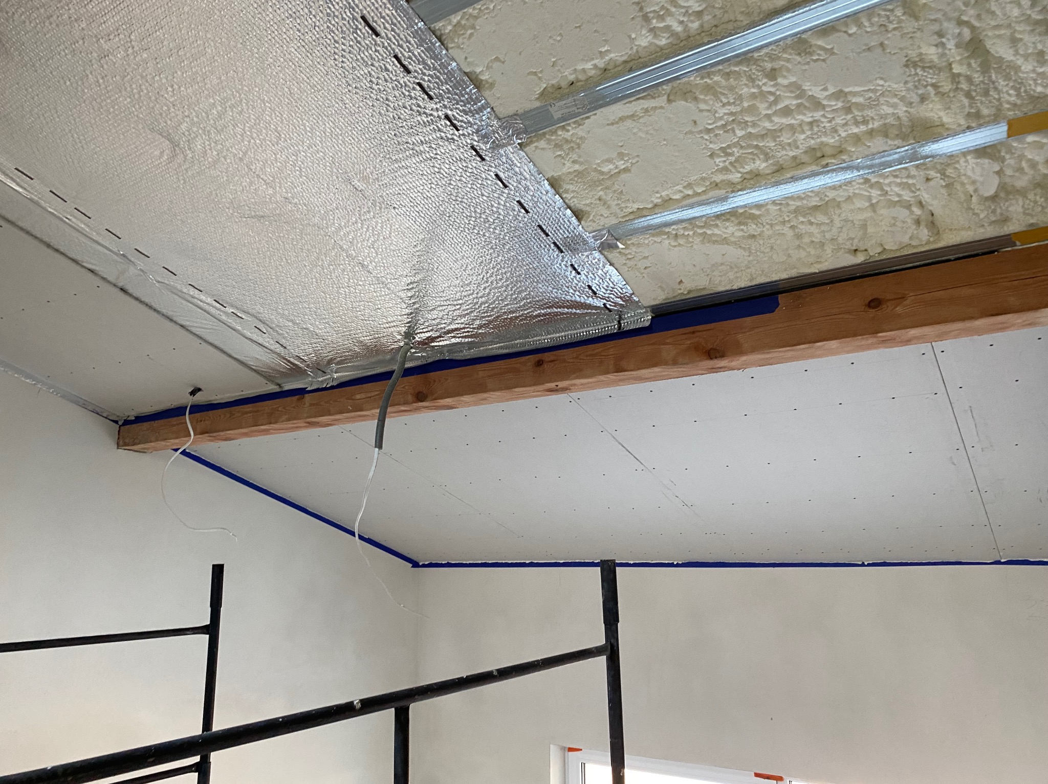 Montaż sufitu podwieszanego: widoczne profile metalowe, izolacja z pianki oraz częściowo zamontowana płyta karton gips z folią aluminiową, zabezpieczona niebieską taśmą malarską przy ścianach.