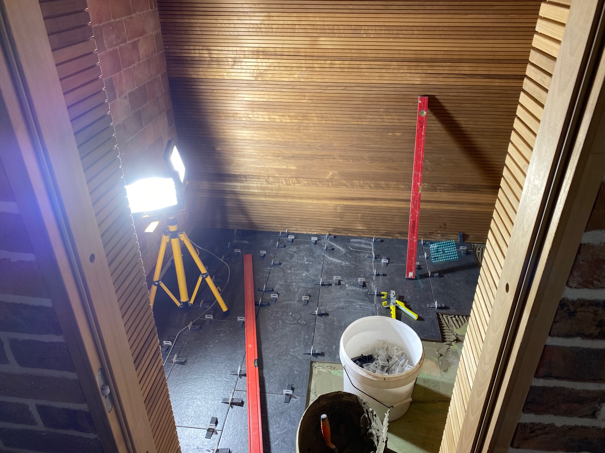 Ukośne ujęcie poddasza z drewnianą boazerią na suficie podczas układania ciemnych płytek podłogowych, widoczne narzędzia, poziomica, wiadro z krzyżykami dystansowymi i lampa na statywie.