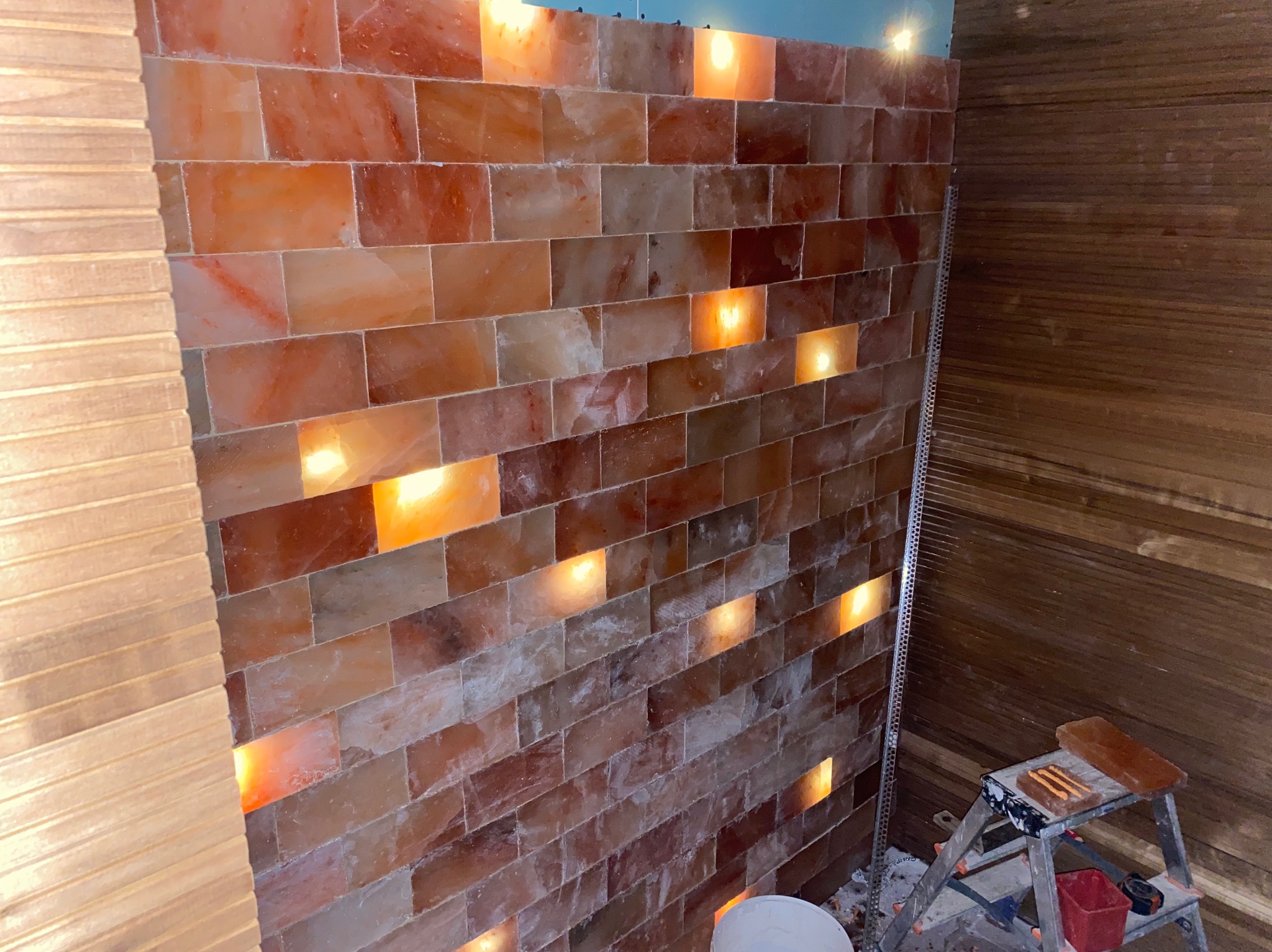 Ściana z bloków soli himalajskiej z podświetleniem, widoczne elementy drewnianej boazerii, drabina malarska i narzędzia w trakcie montażu.