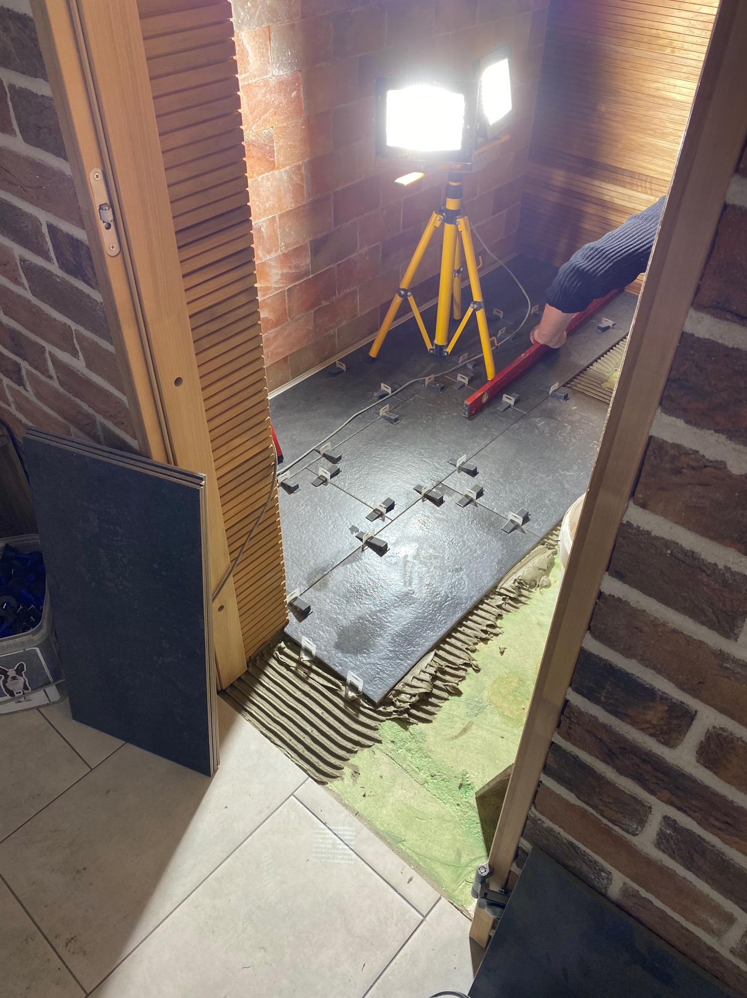 Układanie ciemnych płytek podłogowych w pomieszczeniu z ceglanymi ścianami, widoczne krzyżyki dystansowe, poziomica i lampa na statywie.