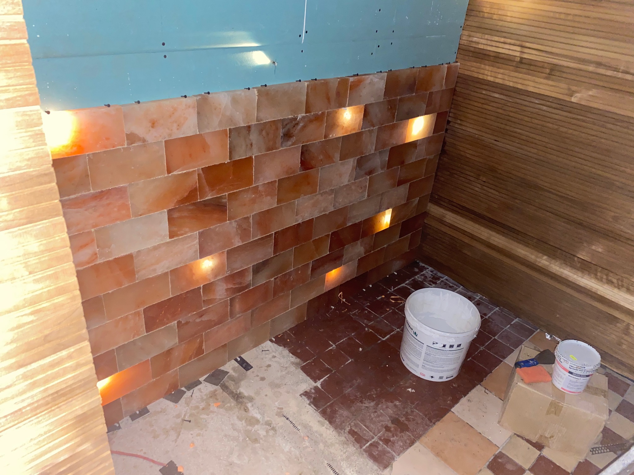 Montaż ściany z cegieł solnych z podświetleniem na płycie karton-gips w trakcie prac wykończeniowych, widoczne wiaderka z farbą i narzędzia na podłodze z częściowo usuniętymi płytkami.