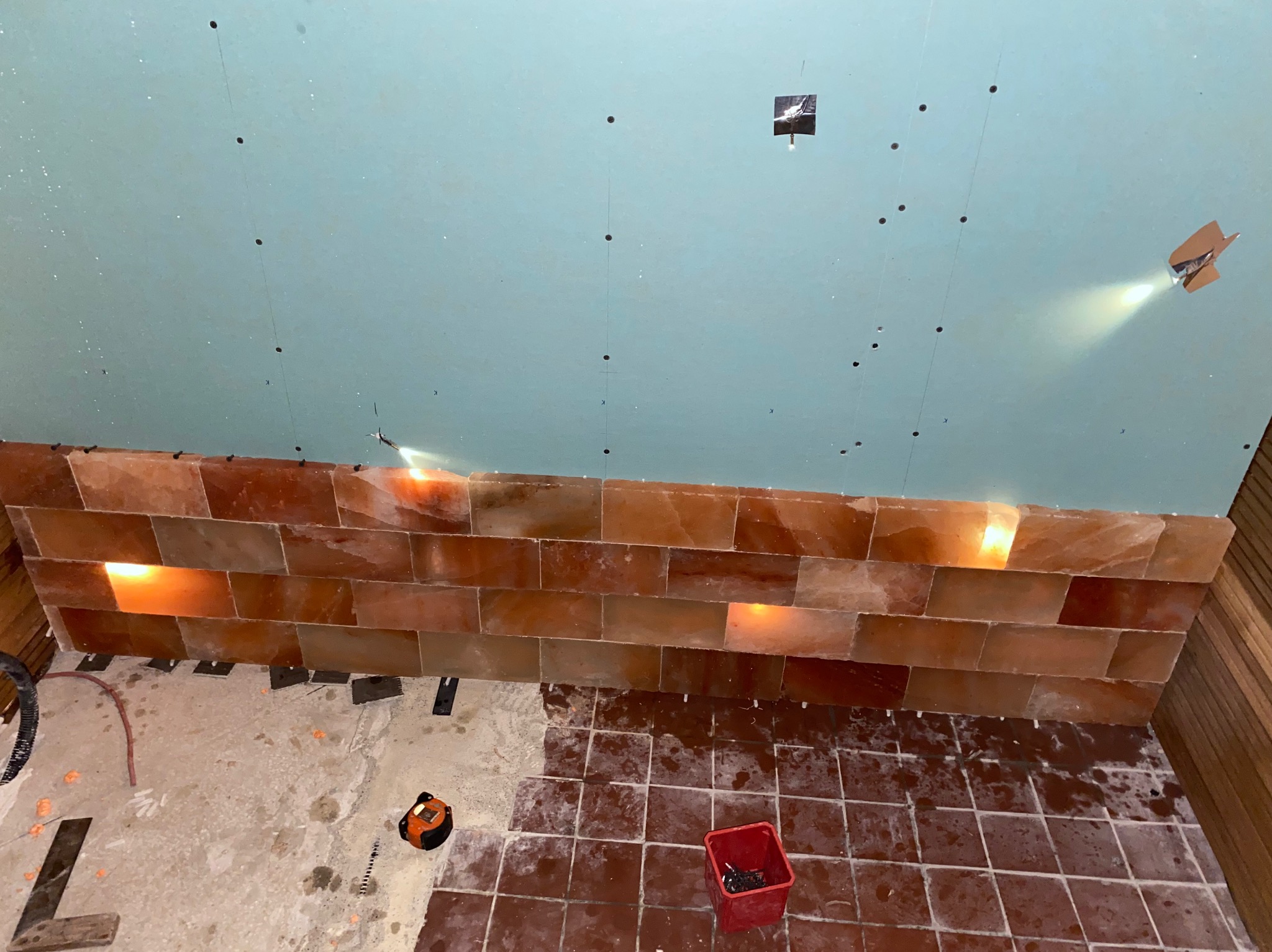 Montaż ściany z cegieł solnych w łazience, widoczne podświetlenie, sufit z płyt kartonowo-gipsowych z zaznaczonymi punktami, częściowo ułożona podłoga z brązowych płytek.
