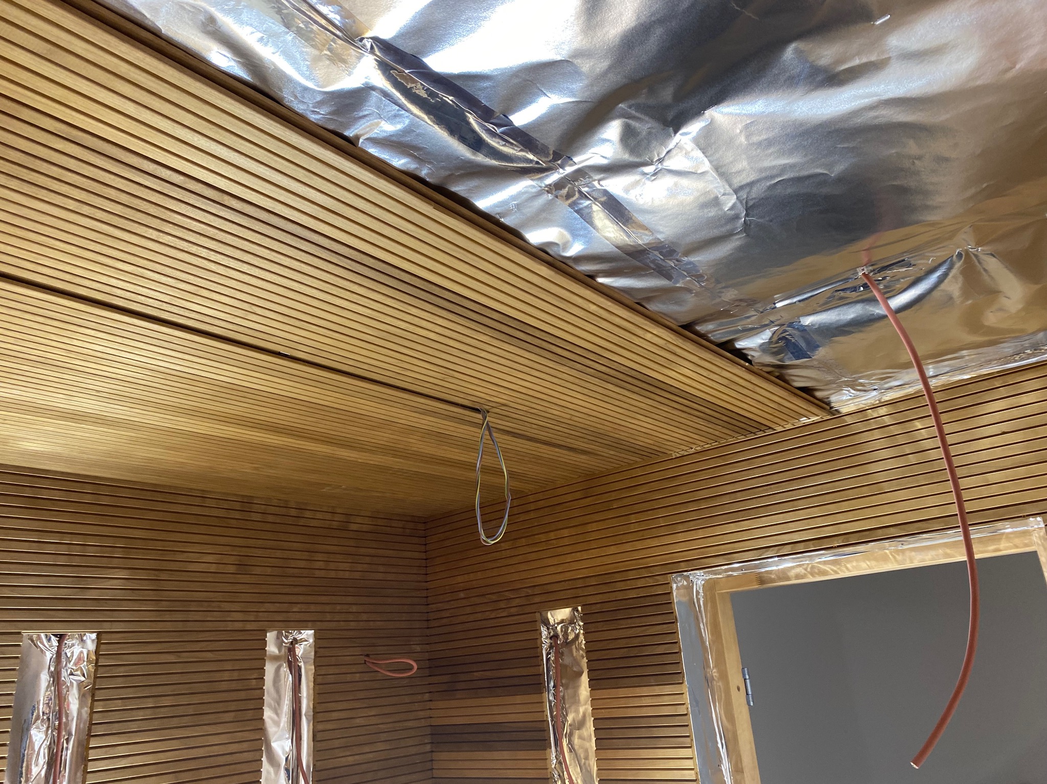 Ściana i sufit wykończone drewnianymi panelami boazeryjnymi w trakcie prac wykończeniowych, widoczne przewody elektryczne i folia aluminiowa izolacyjna.