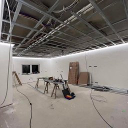 Dany-Bud - Wnętrze w trakcie remontu z widoczną metalową konstrukcją sufitu podwieszanego z zamontowanym oświetleniem LED, narzędzia na drewnianych kozłach, przewody elektryczne, drabina oparta o ścianę.