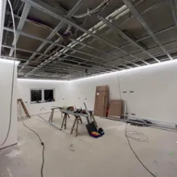 Wnętrze w trakcie remontu z widoczną metalową konstrukcją sufitu podwieszanego z zamontowanym oświetleniem LED, narzędzia na drewnianych kozłach, przewody elektryczne, drabina oparta o ścianę.