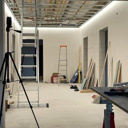 Dany-Bud - Wnętrze w trakcie prac wykończeniowych: widoczna metalowa konstrukcja sufitu podwieszanego, drabina, statyw aparatu i narzędzia na stole warsztatowym.