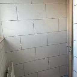 Ściana wyłożona białymi, prostokątnymi płytkami imitującymi marmur, z ciemnymi fugami. Widoczny fragment grzejnika i drzwi z metalową klamką.