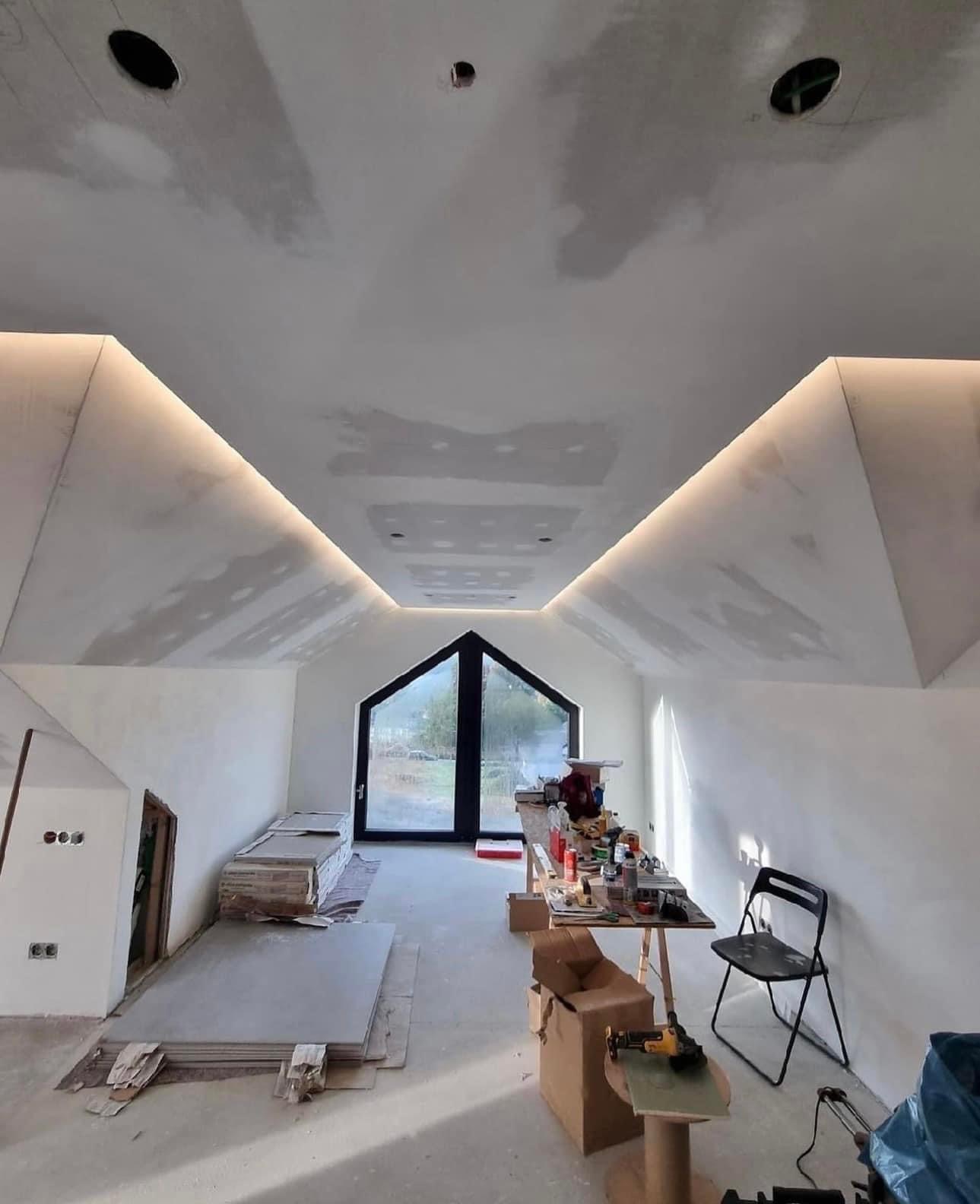 Poddasze w trakcie prac wykończeniowych: zamontowane płyty karton-gips na skosach i suficie, widoczne otwory na oświetlenie, listwy LED, okno w kształcie trapezu, narzędzia i materiały budowlane...