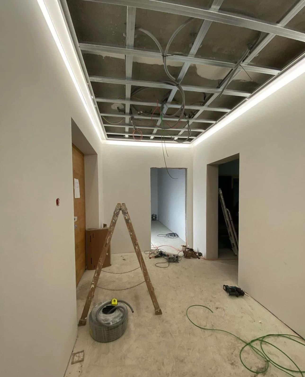 Wnętrze w trakcie remontu: szkielet sufitu podwieszanego z oświetleniem LED, drabina oparta o ścianę, widoczne kable i narzędzia na podłodze.