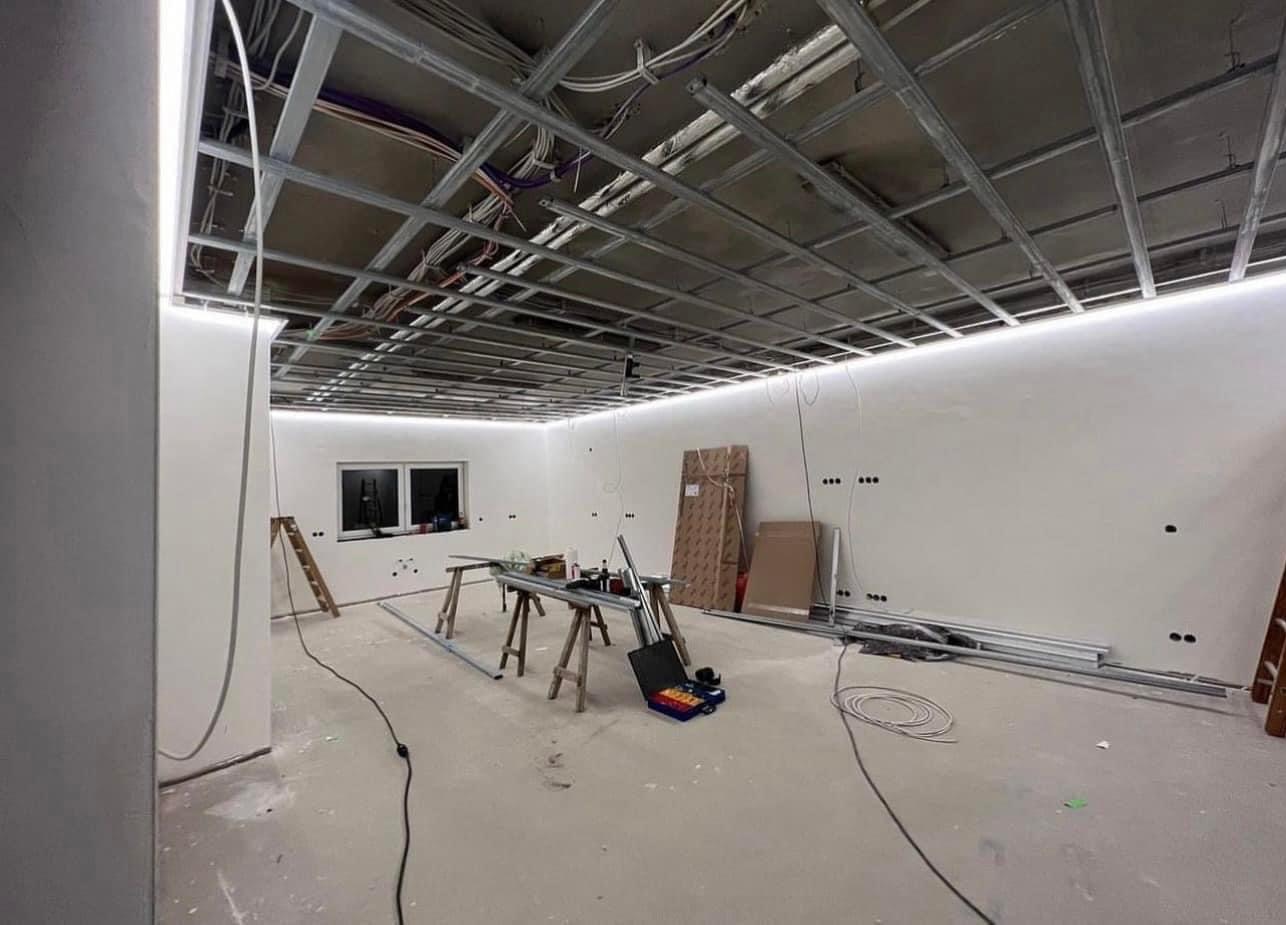 Wnętrze w trakcie remontu z widoczną metalową konstrukcją sufitu podwieszanego z zamontowanym oświetleniem LED, narzędzia na drewnianych kozłach, przewody elektryczne, drabina oparta o ścianę.