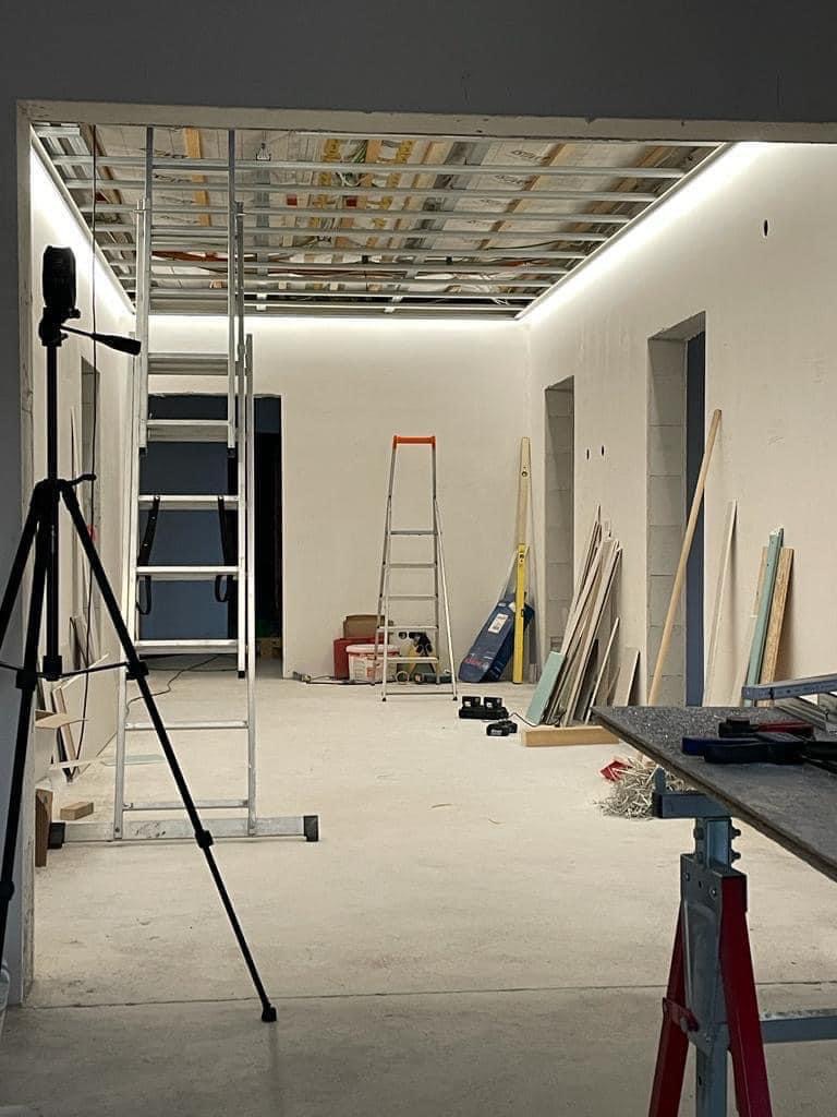 Wnętrze w trakcie prac wykończeniowych: widoczna metalowa konstrukcja sufitu podwieszanego, drabina, statyw aparatu i narzędzia na stole warsztatowym.