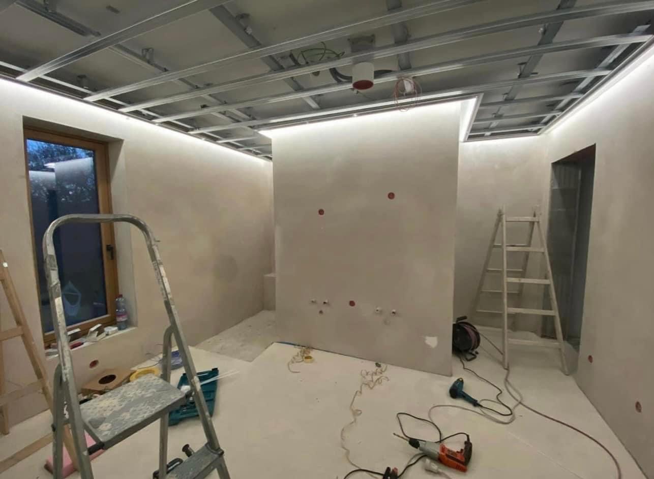 Wnętrze w trakcie prac wykończeniowych: zamontowana metalowa konstrukcja sufitu podwieszanego z oświetleniem LED, ściany po wstępnym nałożeniu gładzi, widoczne narzędzia i drabiny.
