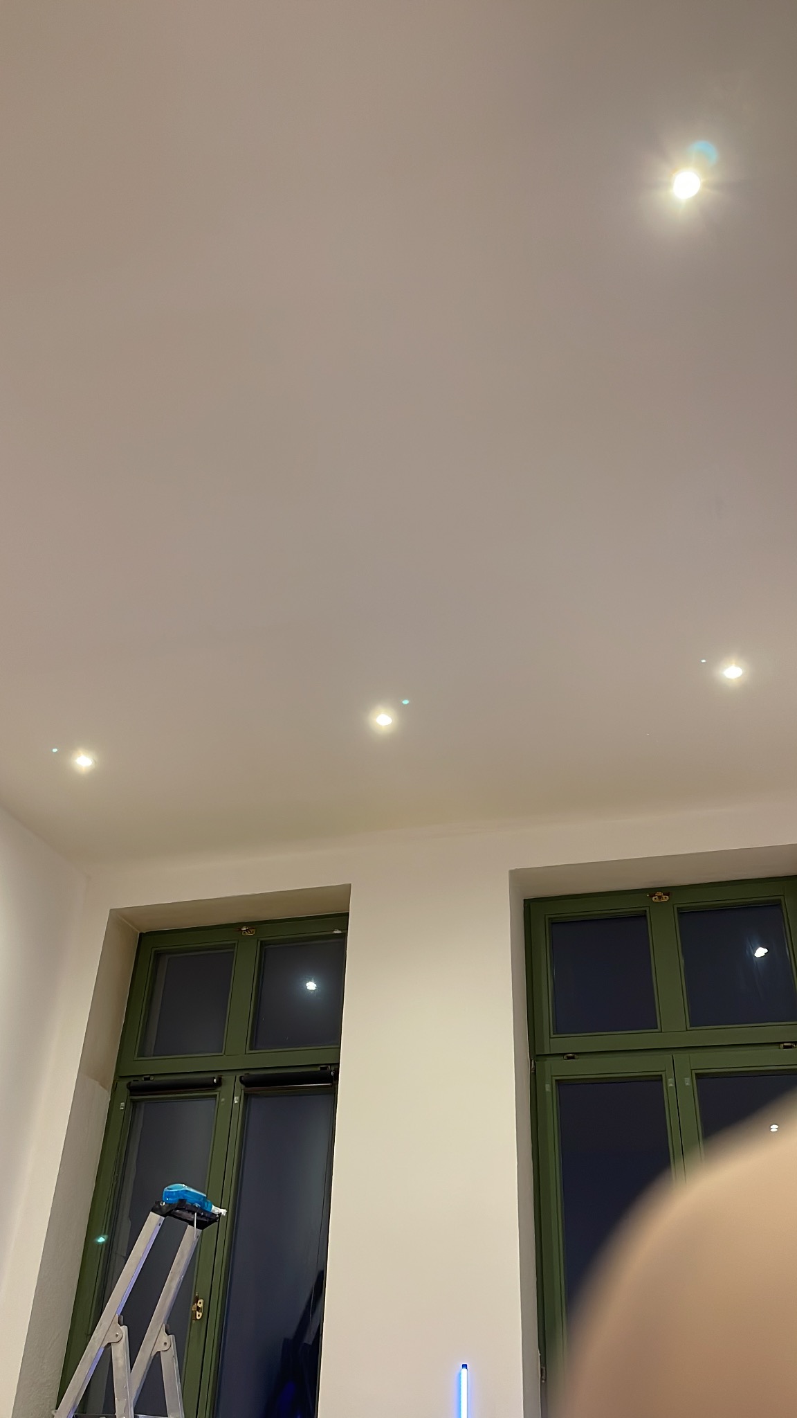 Wnętrze z dwoma oknami o zielonych ramach, białą ścianą i sufitem z trzema punktowymi lampami; drabina oparta o ścianę przy oknie, widoczny fragment ciała w prawym dolnym rogu.