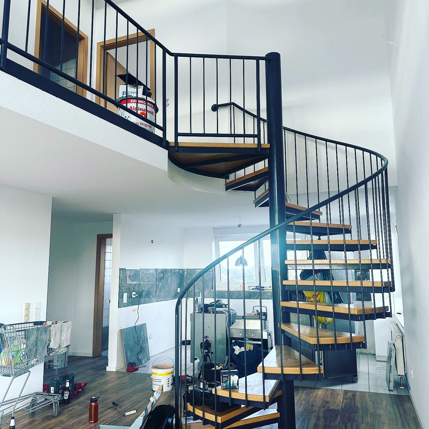 Wnętrze w trakcie remontu z drewnianymi schodami spiralnymi z czarną metalową balustradą, widoczne narzędzia i materiały budowlane, podłoga z paneli, fragment ściany z niedokończonymi płytkami.