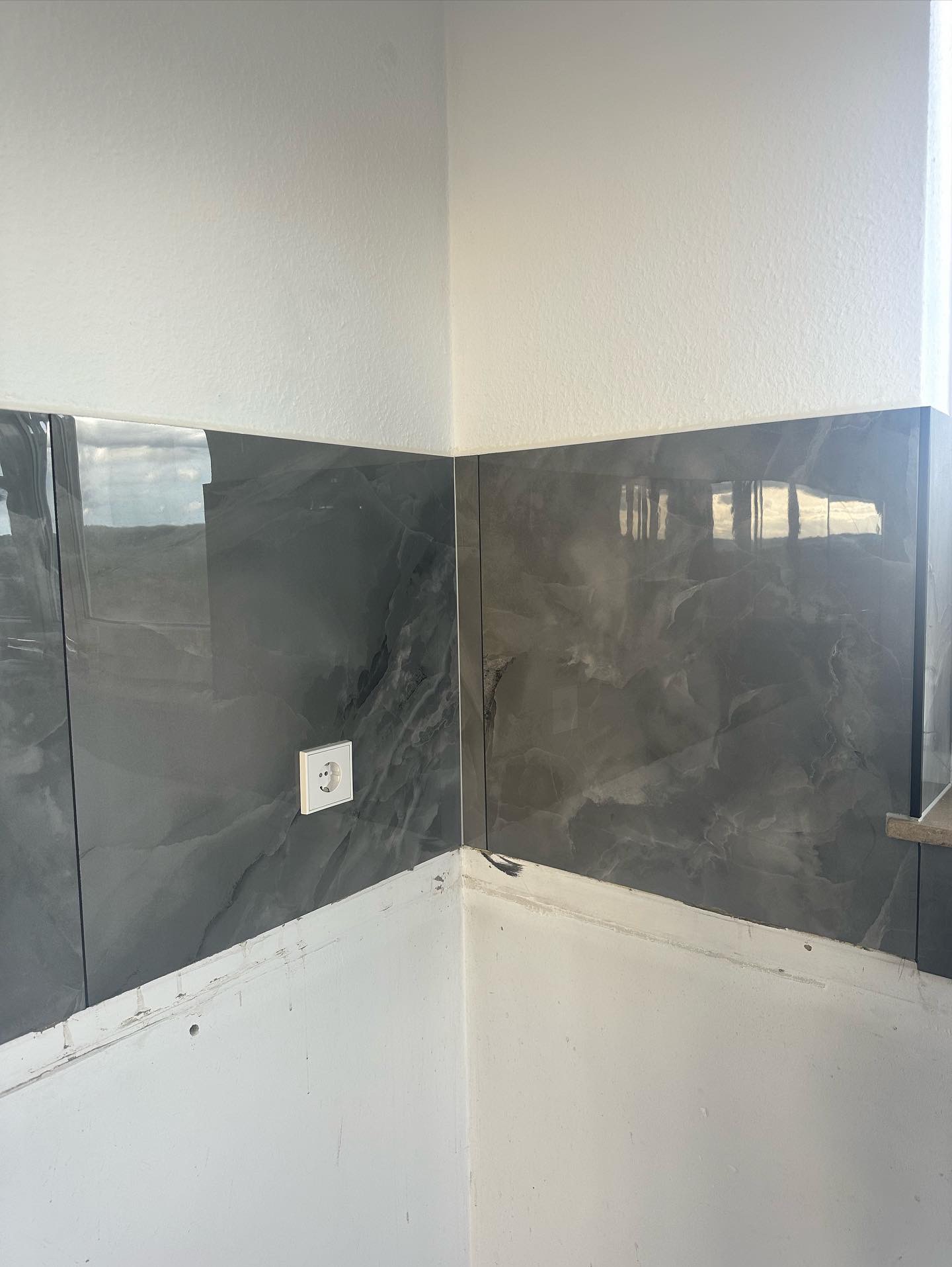 Ściana w rogu pomieszczenia z częściowo położonymi szarymi płytkami imitującymi marmur, widoczne gniazdko elektryczne i niedokończone wykończenie powyżej płytek.
