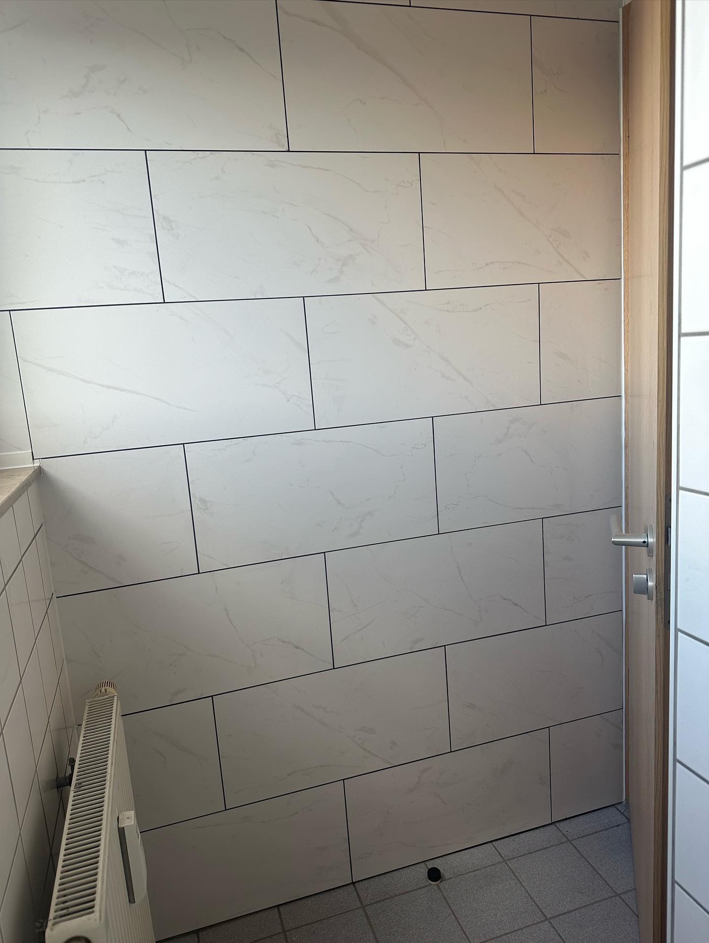 Ściana wyłożona białymi, prostokątnymi płytkami imitującymi marmur, z ciemnymi fugami. Widoczny fragment grzejnika i drzwi z metalową klamką.