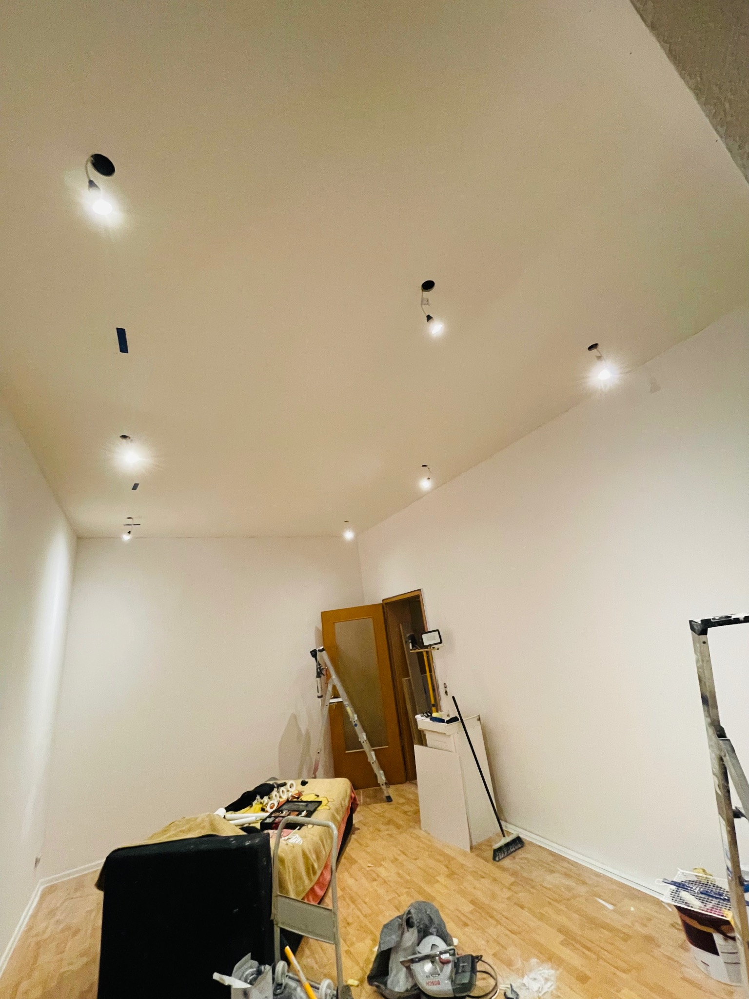 Pokój w trakcie remontu: świeżo pomalowane ściany na biało, widoczne punkty oświetleniowe w suficie, zabezpieczone taśmą, podłoga parkietowa, drabina oparta o ścianę, narzędzia malarskie...