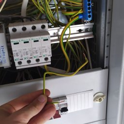 ElektroInstalAC Jarosław Żołneczko - Ręka elektryka podłącza przewód ochronny (żółto-zielony) do ogranicznika przepięć w skrzynce elektrycznej. Widoczne bezpieczniki, listwa zaciskowa i inne przewody.