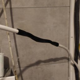 ElektroInstalAC Jarosław Żołneczko - Połączenie przewodów elektrycznych zabezpieczone czarną termokurczliwą izolacją na tle szarej ściany z płytek.