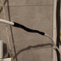 ElektroInstalAC Jarosław Żołneczko - Połączenie kabli elektrycznych zabezpieczone czarną taśmą izolacyjną na tle szarej ściany z płytek ceramicznych.