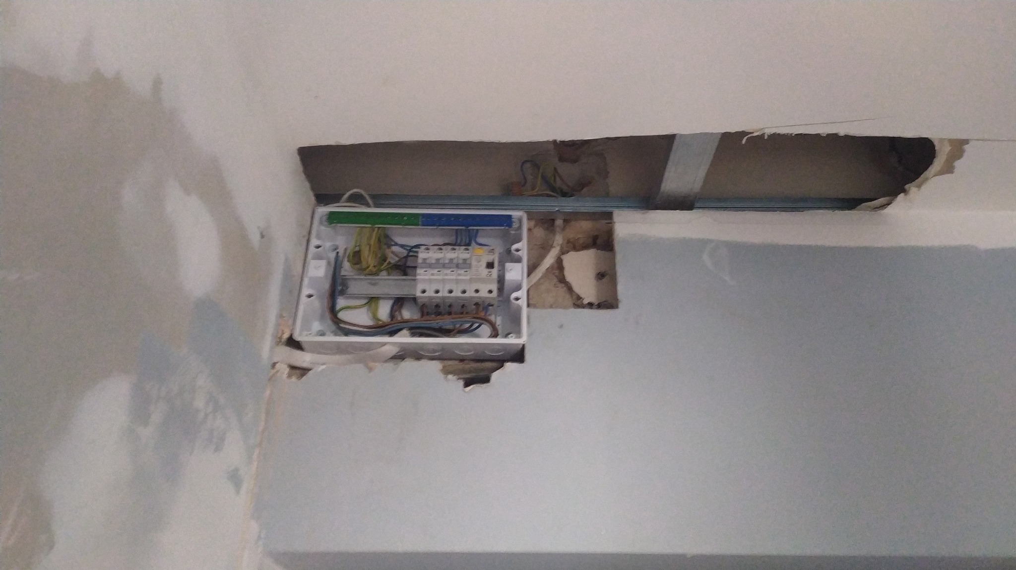 Otwarta skrzynka bezpiecznikowa z widocznymi przewodami i bezpiecznikami, zamontowana w ścianie w trakcie remontu, z widocznymi niedoskonałościami wokół otworu montażowego.