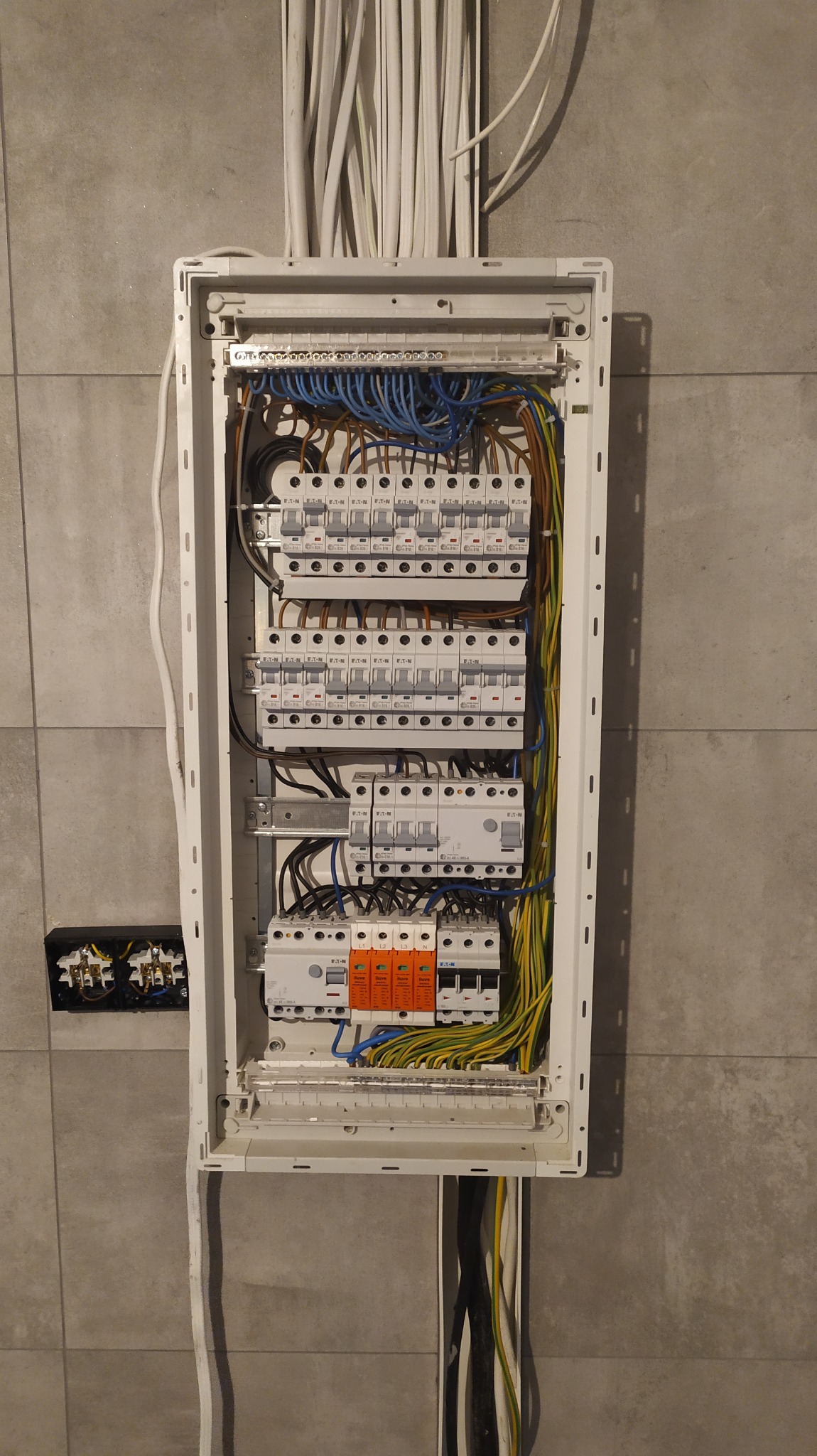 Wnętrze rozdzielni elektrycznej z zamontowanymi bezpiecznikami, ogranicznikami przepięć i okablowaniem w kolorach: niebieskim, brązowym, żółto-zielonym, zamontowanej na szarej ścianie z płytek.