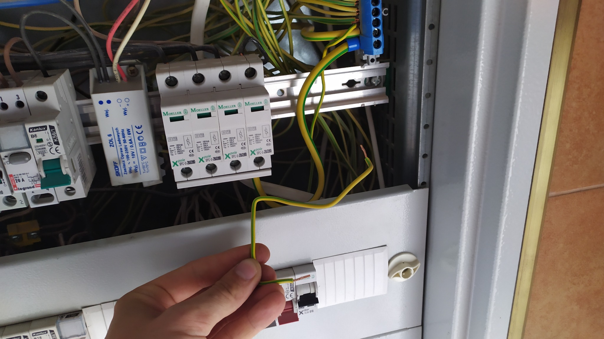 Ręka elektryka podłącza przewód ochronny (żółto-zielony) do ogranicznika przepięć w skrzynce elektrycznej. Widoczne bezpieczniki, listwa zaciskowa i inne przewody.