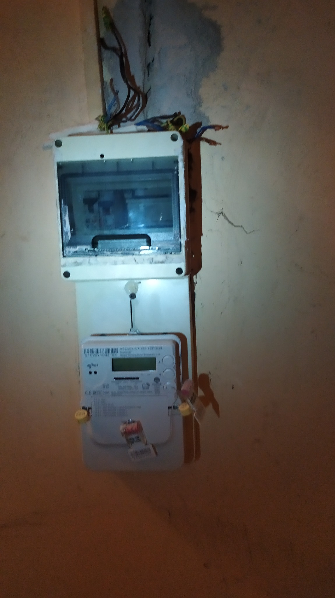 Zdjęcie licznika elektrycznego z otwartą skrzynką bezpiecznikową, widoczne przewody i uszkodzona ściana, sugerujące interwencję elektryka w celu usunięcia nielegalnego poboru energii.