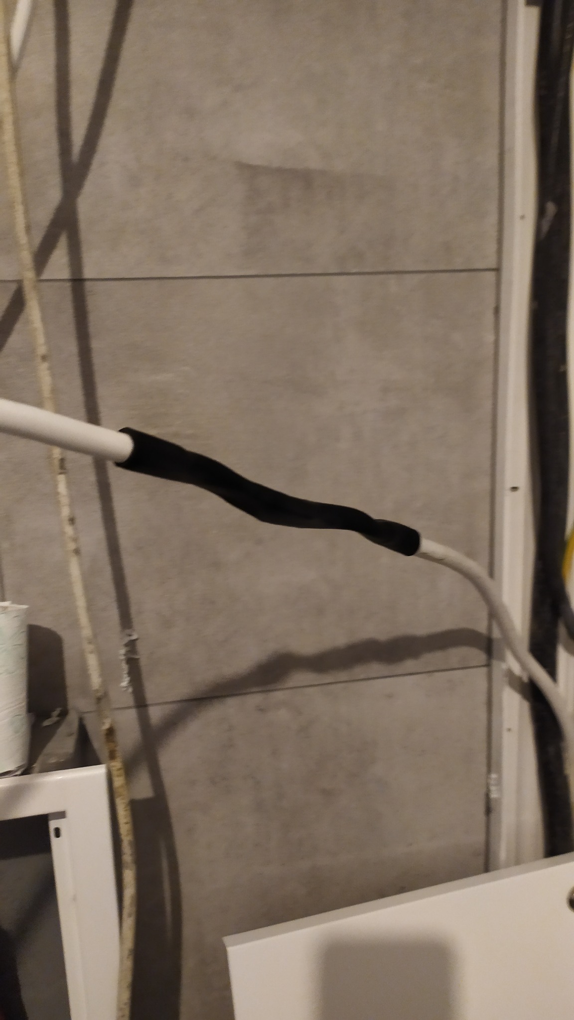 Połączenie kabli elektrycznych zabezpieczone czarną taśmą izolacyjną na tle szarej ściany z płytek ceramicznych.