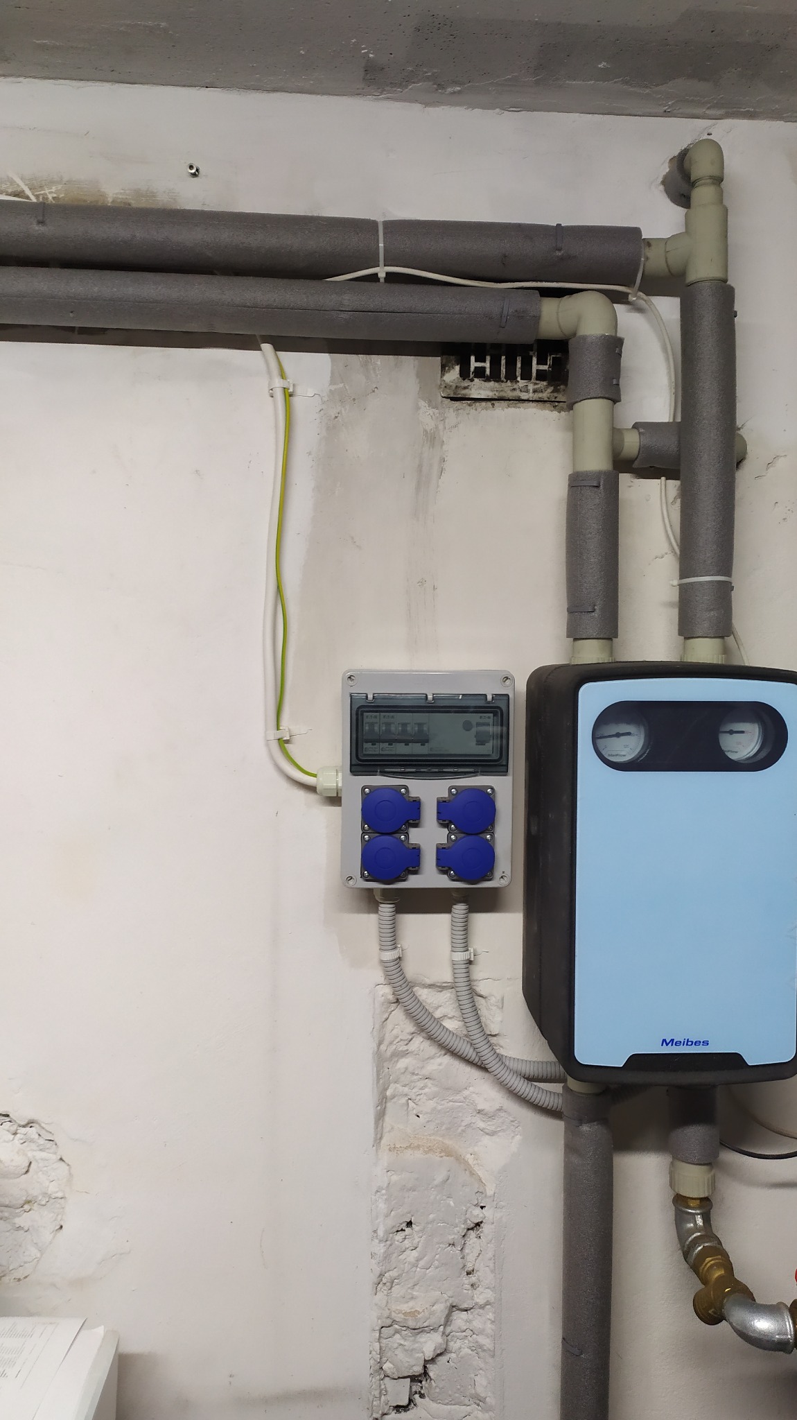 Szara rozdzielnica elektryczna z czterema niebieskimi gniazdami, umieszczona na białej ścianie obok jasnoniebieskiego urządzenia Meibes z dwoma okrągłymi wskaźnikami, rury w szarej izolacji...