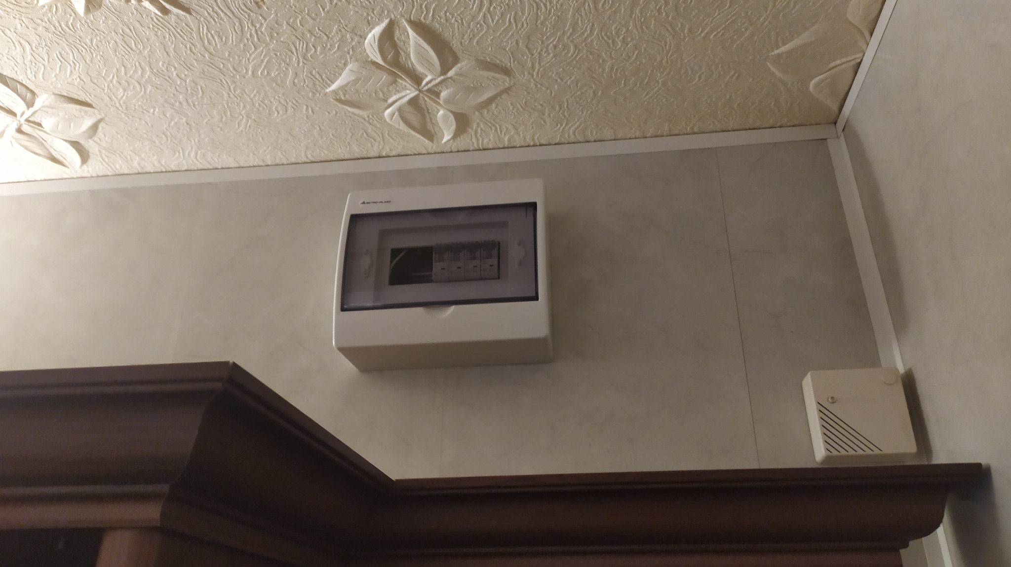 Zamknięta, biała skrzynka z wyłącznikami nadprądowymi zamontowana na ścianie nad ciemnobrązową framugą drzwiową, widoczny również mały biały moduł alarmowy, na tle ściany z jasnych paneli...