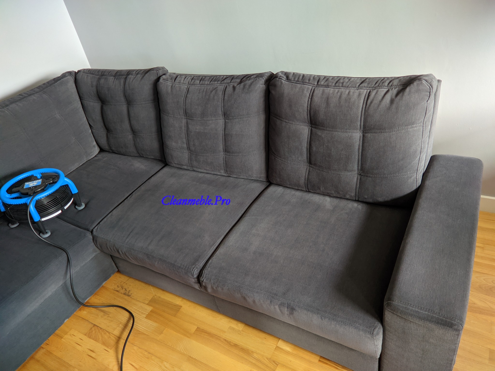 Szara, narożna kanapa z kwadratowymi przeszyciami na poduszkach, obok niebieskie urządzenie do czyszczenia tapicerki, widoczny kabel zasilający.