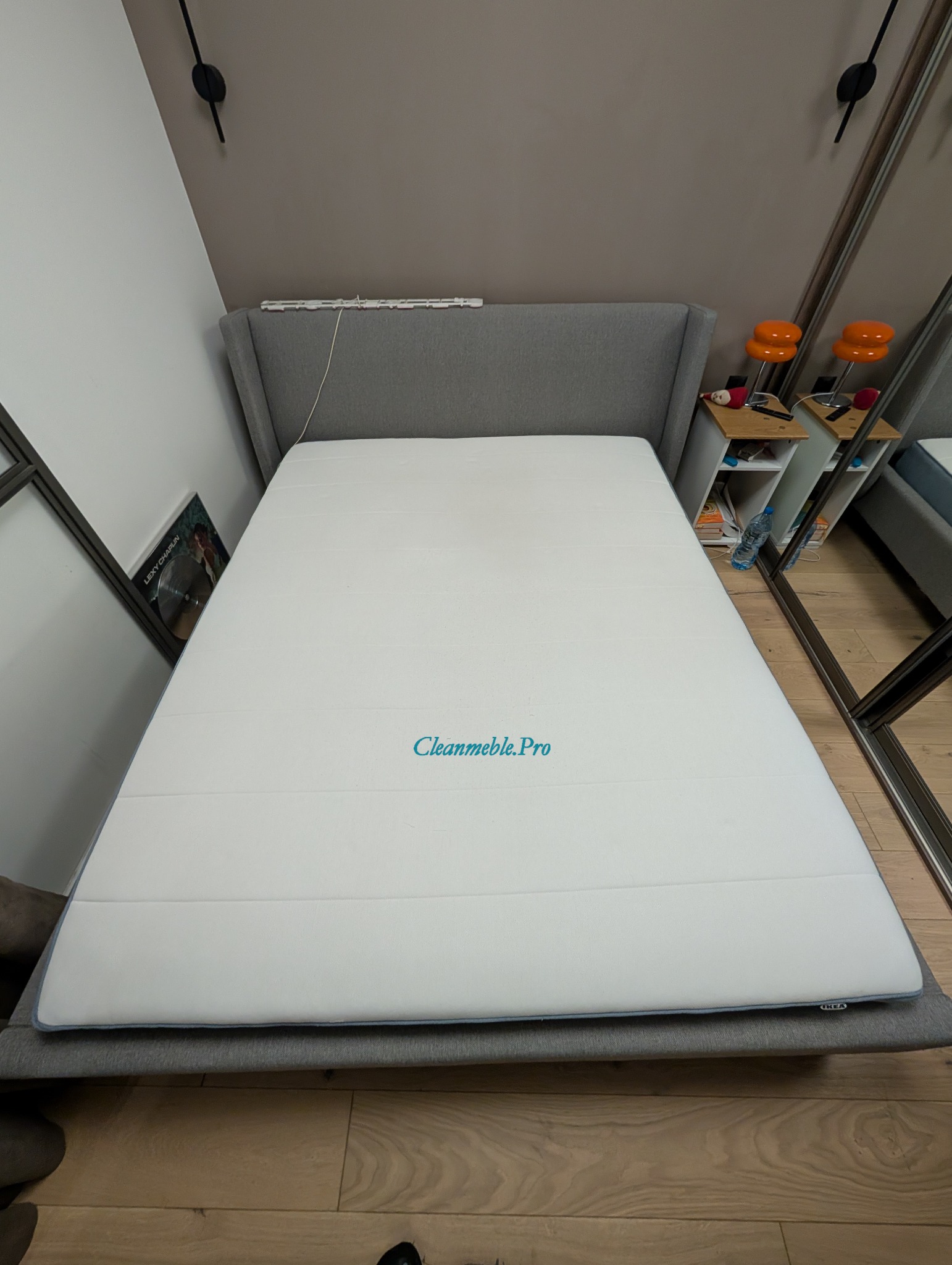 Biały materac na szarym łóżku z zagłówkiem, widok z góry. Na materacu widnieje napis 'Cleanmeble.Pro'. W tle elementy wystroju wnętrza.