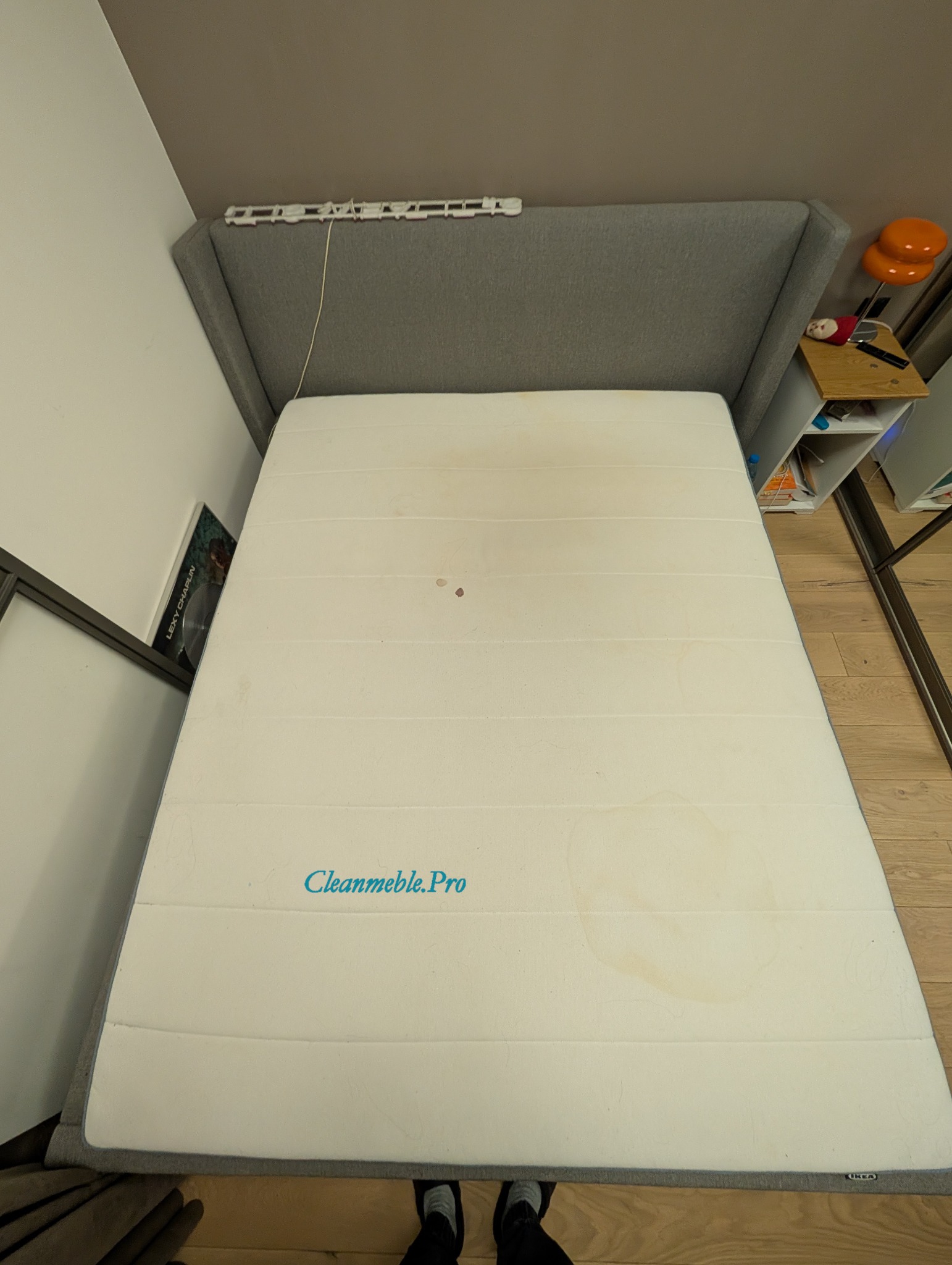 Materac z widocznymi plamami, leżący na łóżku z szarą ramą. Na materacu widoczny napis Cleanmeble.Pro. Ujęcie z góry, widoczne fragmenty podłogi i stóp.