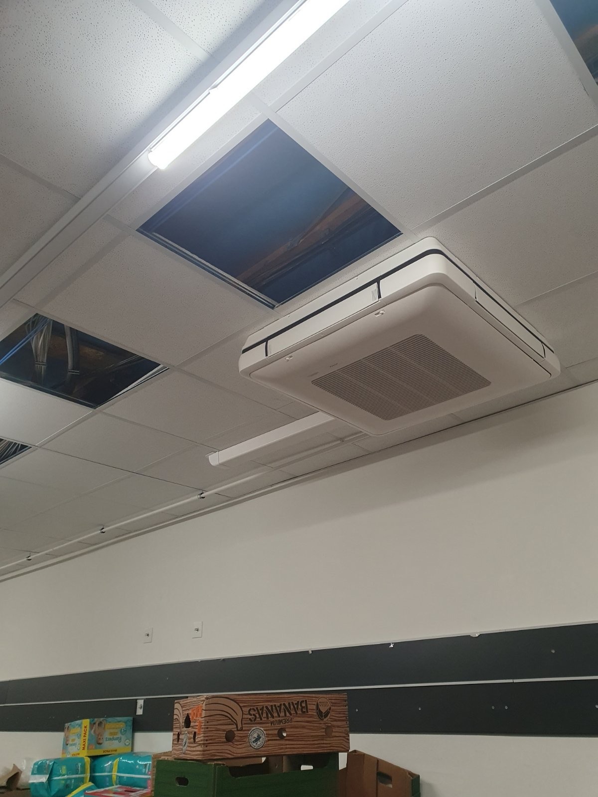 Kasetonowa jednostka klimatyzacyjna zamontowana w suficie podwieszanym biura, widoczne puste przestrzenie po brakujących płytach sufitowych oraz kartony na dole ściany.