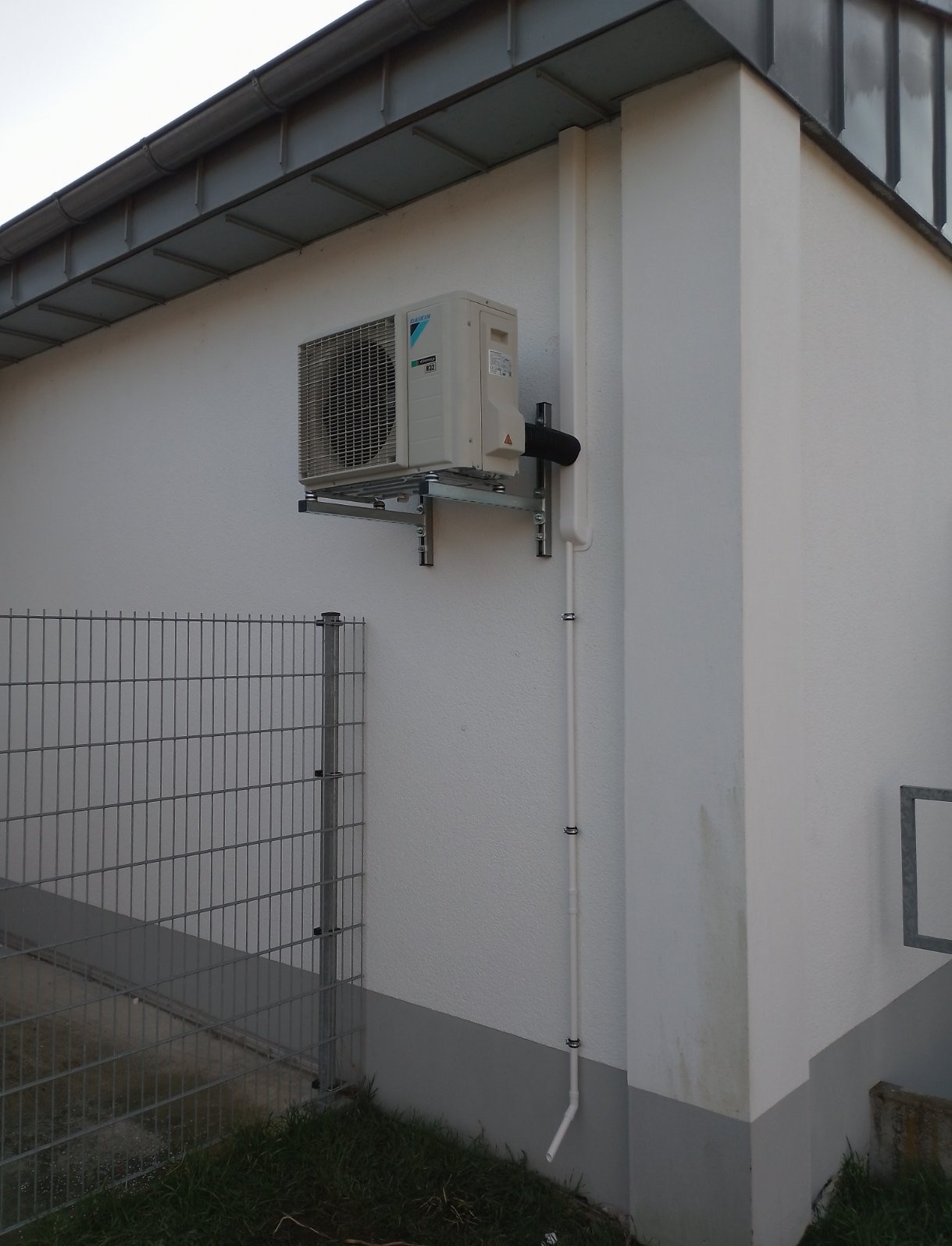 Zewnętrzna jednostka klimatyzacji Daikin zamontowana na metalowym wsporniku na ścianie budynku z widoczną instalacją odprowadzającą skropliny.