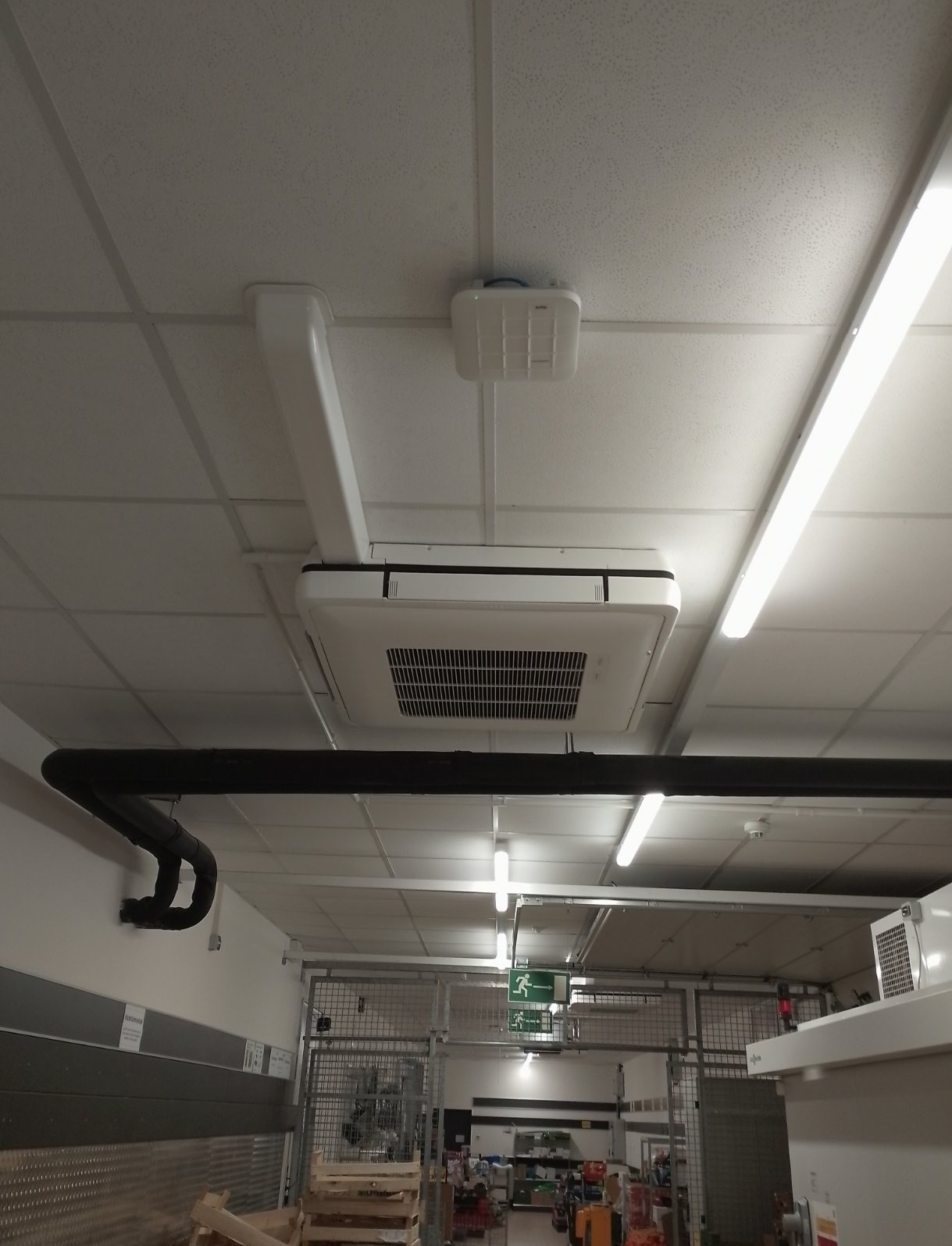 Sufitowa jednostka klimatyzacyjna kasetonowa w przestrzeni handlowej, widoczne rury instalacyjne i oświetlenie sufitowe.