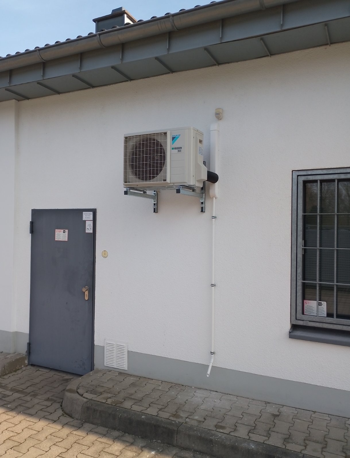 Zewnętrzna jednostka klimatyzacji Daikin zamontowana na metalowych wspornikach na białej ścianie budynku, z widoczną instalacją odprowadzającą skropliny, obok szare drzwi i okno z kratą.