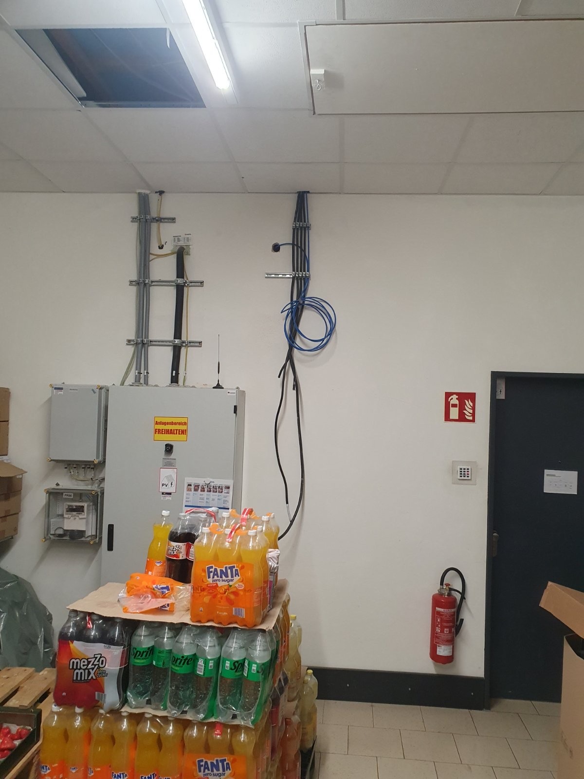 Instalacja elektryczna w pomieszczeniu biurowym z widocznym okablowaniem, skrzynką elektryczną i stosem napojów gazowanych na palecie.