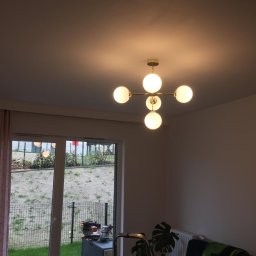 MULTI MONTI Janusz Zduńczyk - Nowoczesny żyrandol z pięcioma mlecznymi kulami zawieszony w pokoju, widok na zewnątrz przez okno, drabina oparta o ścianę, narzędzia widoczne na zewnątrz okna.