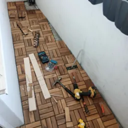 Montaż drewnianych paneli podłogowych na balkonie, widoczne narzędzia: wiertarka, młotek, miarka, skrzynki z wiertłami, deski do docinania.