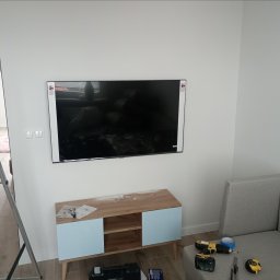 MULTI MONTI Janusz Zduńczyk - Telewizor zawieszony na białej ścianie nad komodą z błękitnymi drzwiczkami. Obok leży szara kanapa, a na niej wiertarka, taśma malarska i miarka. Na komodzie widoczne akcesoria montażowe, a po...