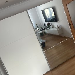 MULTI MONTI Janusz Zduńczyk - Nowoczesna szafa przesuwna z białymi frontami i lustrami, odbijająca wnętrze pokoju z telewizorem i drewnianą podłogą.