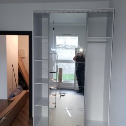 MULTI MONTI Janusz Zduńczyk - Wnętrze pokoju w trakcie wykańczania: biała szafa z przesuwnymi lustrzanymi drzwiami, drabina oparta o okno, kartony po meblach przy ścianie, odbicie osoby robiącej zdjęcie.